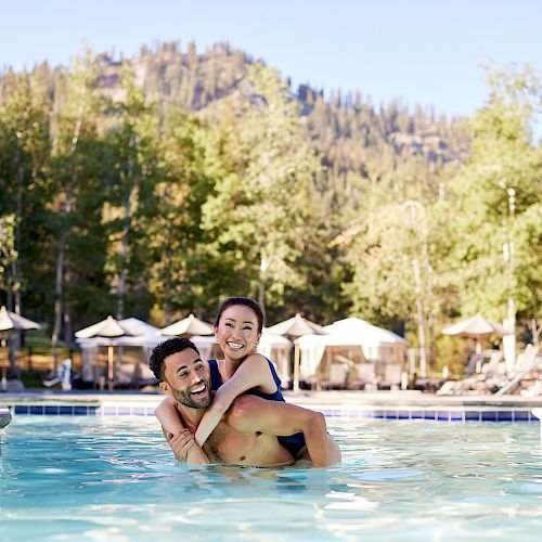 A couple enjoys time together in a pool, with scenic trees and mountains in the background on a sunny day, surrounded by lounge chairs and umbrellas.