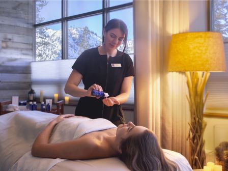 A woman prepares massage oil in a serene spa setting, with another person relaxing on a table, candles lit, and snowy mountains outside.