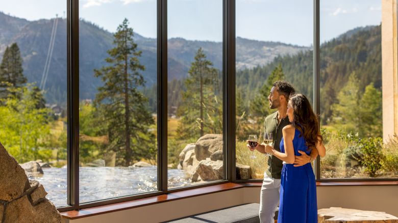 A couple stands indoors, looking out a large window with a scenic view of mountains and trees. They appear to be enjoying a relaxing moment.