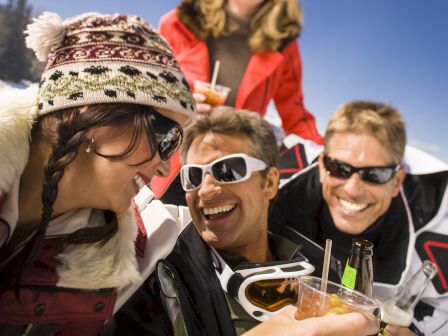 Several people in winter attire are laughing and enjoying drinks while on a snowy slope, creating a lively and fun atmosphere.