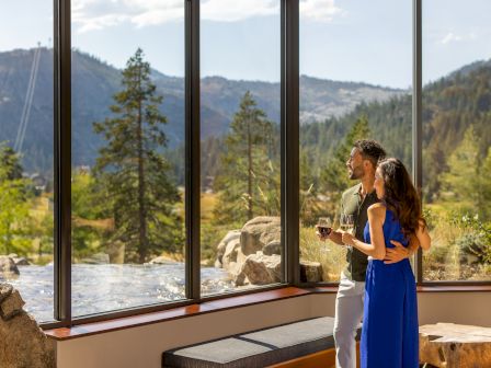 A couple is standing indoors, enjoying a scenic view of mountains and trees through large windows while holding wine glasses.