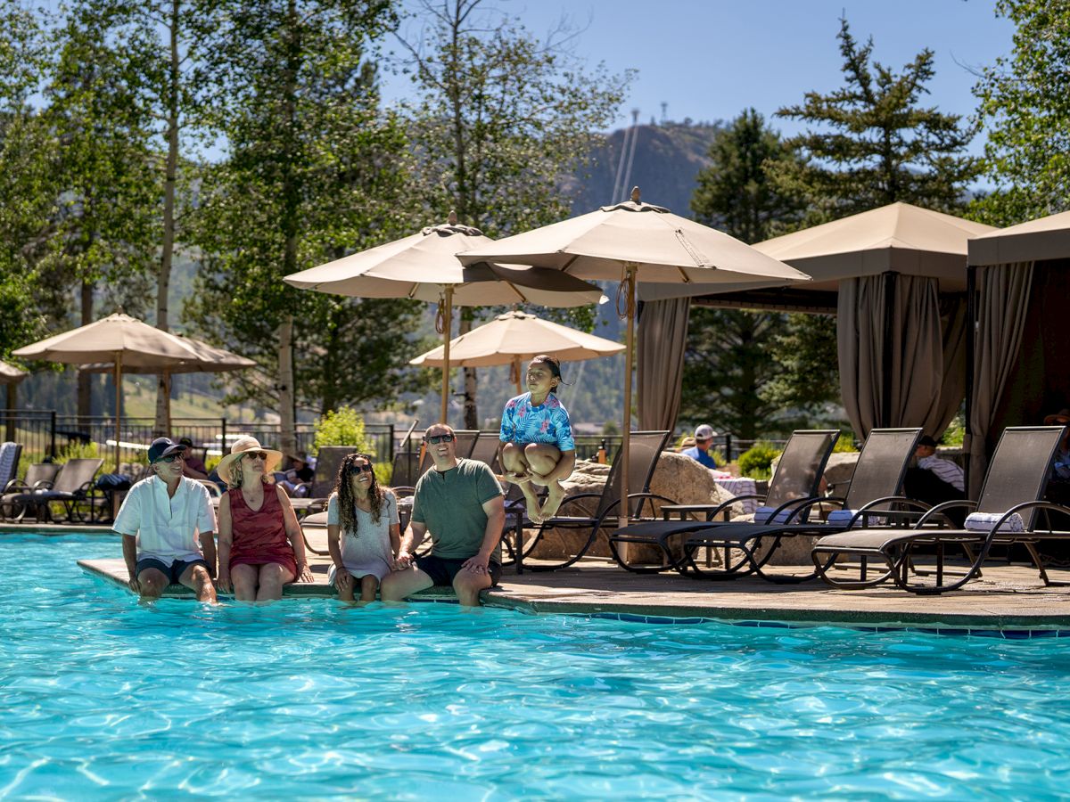 People relaxing at a poolside with lounge chairs, umbrellas, and a cabana area; mountains in the background. Always end with a period.