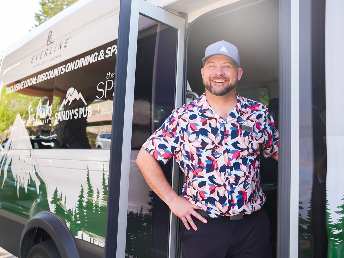 A smiling man in a colorful short-sleeve shirt and cap stands in the doorway of a branded van with forest graphics.