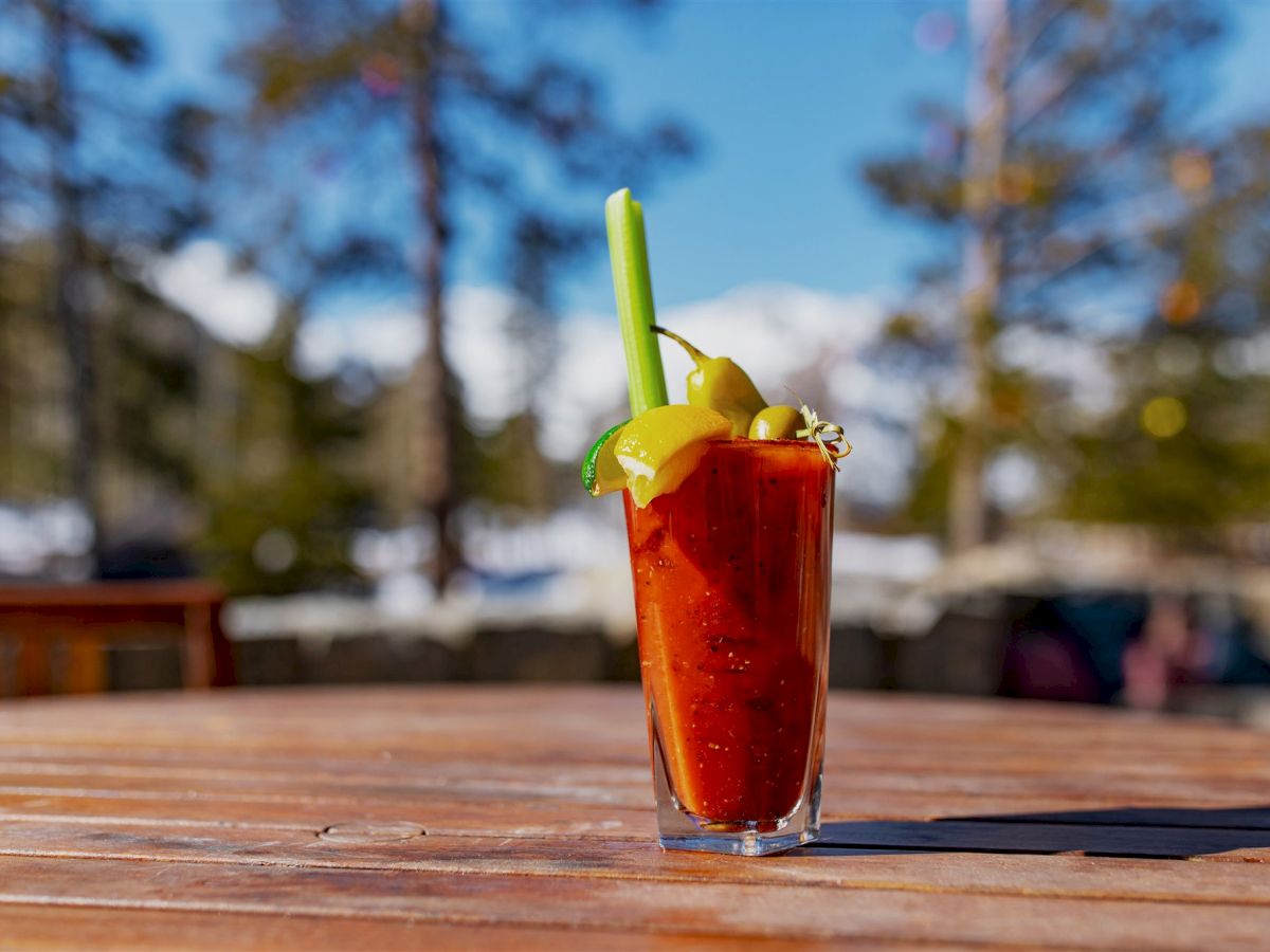 A red cocktail garnished with a celery stick, pickles, olives, lemon slice, and a pepper, placed on a wooden table outdoors.