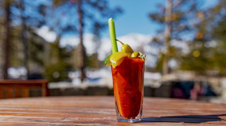 A Bloody Mary cocktail garnished with celery, lemon, and olives sits on a wooden table outdoors, surrounded by a blurred forest background.