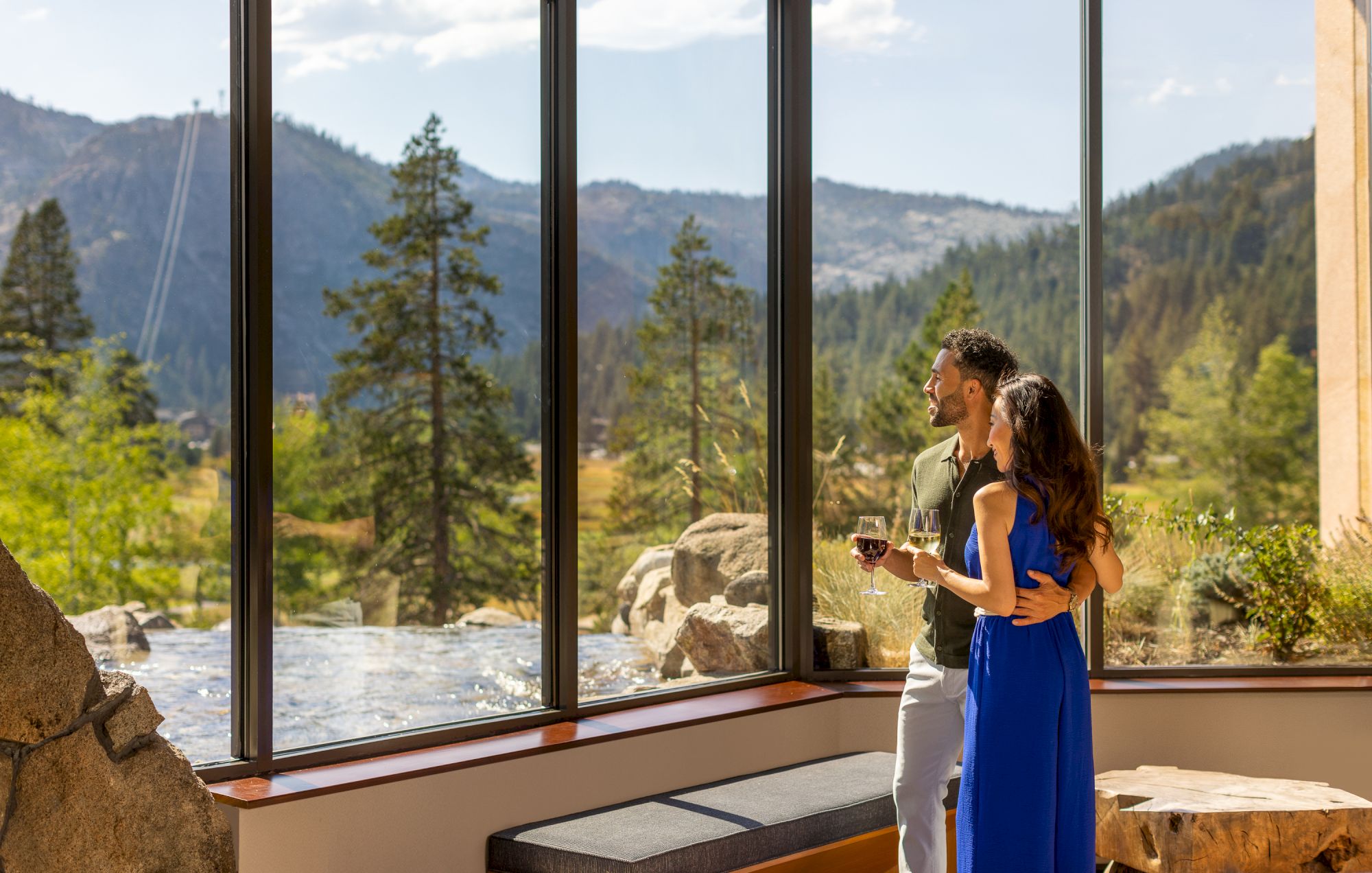 A couple stands indoors by large windows, looking out at a scenic mountain view with trees, holding hands and enjoying a peaceful moment together.