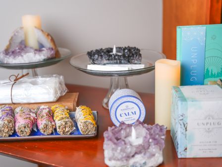 A cozy display of candles, crystals, and colorful treats on a wooden table: cakes, a boxed gift, and a turquoise book beside a purple cluster crystal.