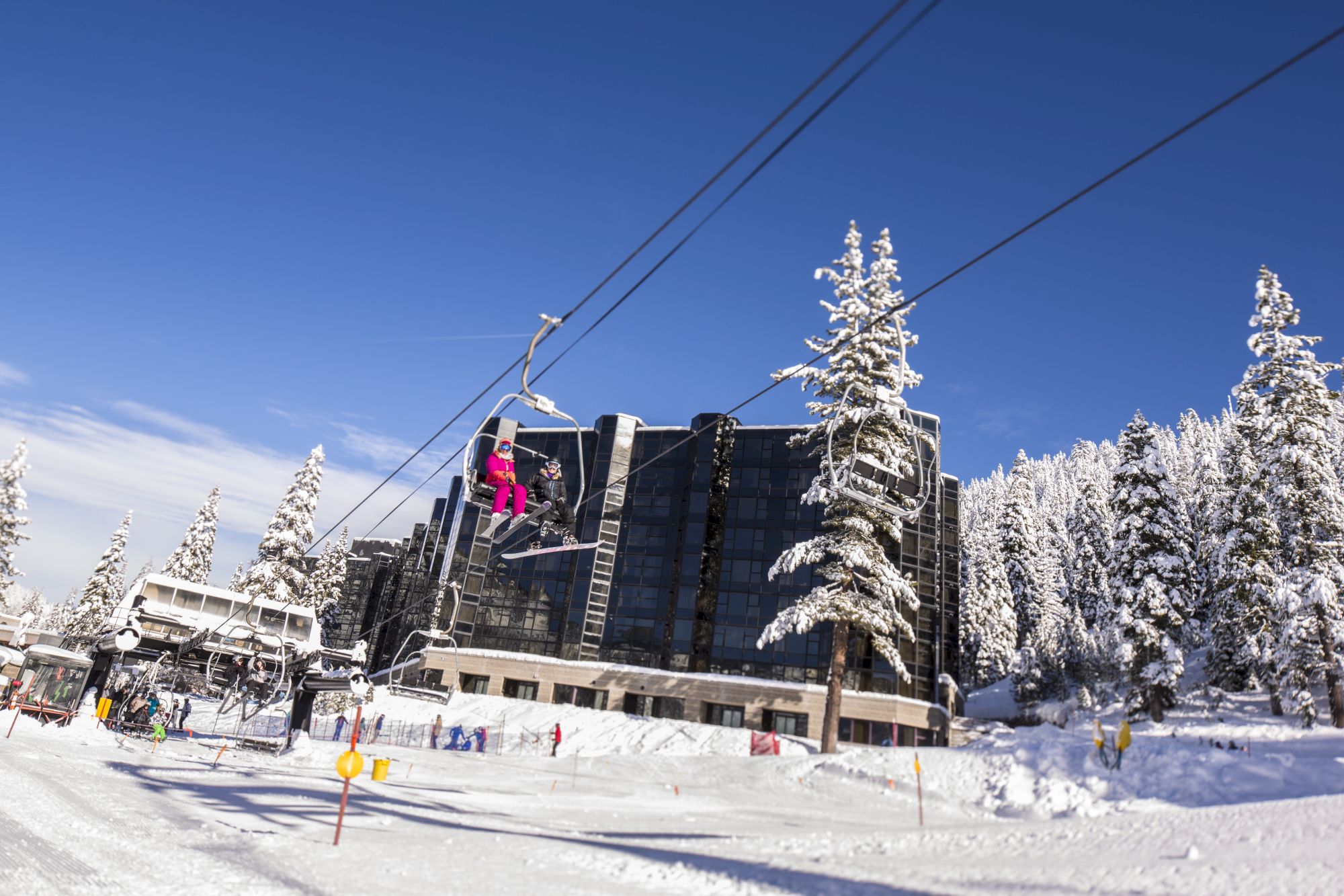 A snowy ski resort with a chairlift transporting people; snow-covered trees and a large building in the background under a clear blue sky.