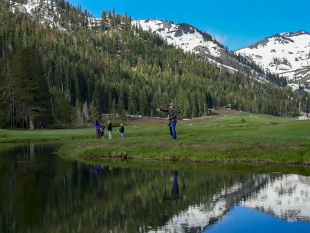 Four people are standing by a reflective pond in a mountainous area with snow-capped peaks and lush green trees in the background.