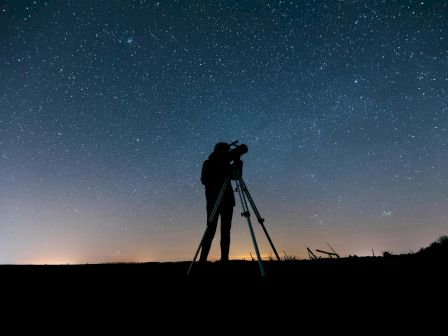 A person is observing the night sky through a telescope, standing under a star-filled sky with a faint glow on the horizon.