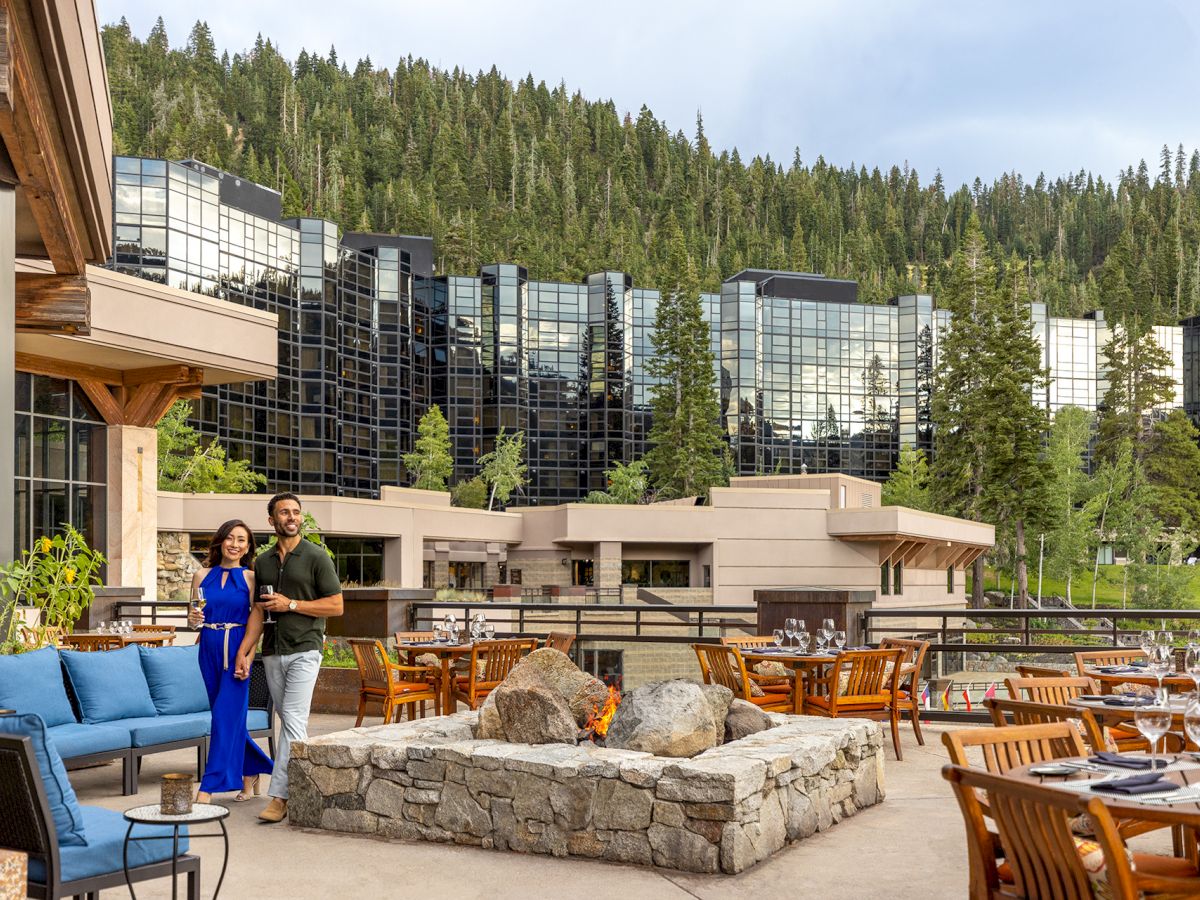 A couple stands by a cozy outdoor seating area with blue couches, a stone fire pit, and wooden tables, in front of a modern glass hotel set among trees.