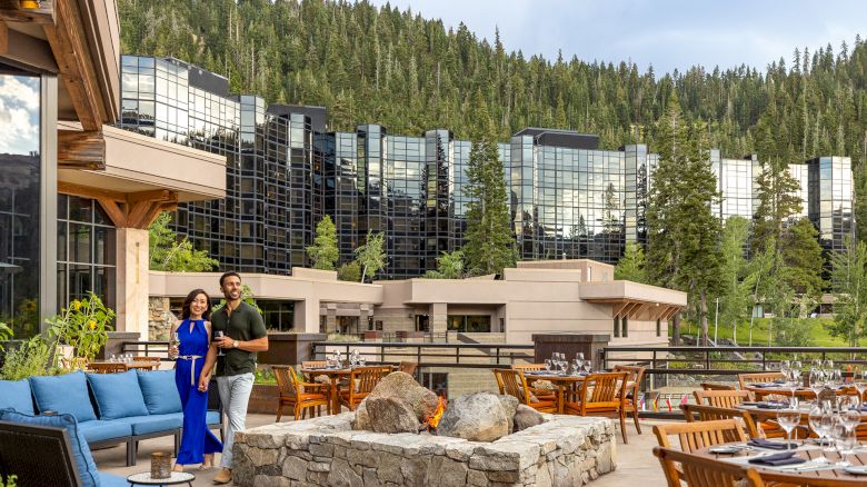 A couple stands by a cozy outdoor seating area with blue couches, a stone fire pit, and wooden tables, in front of a modern glass hotel set among trees.