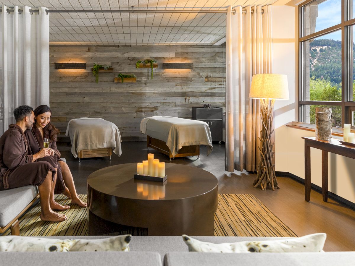 A cozy spa lounge with two people on a sofa, a round coffee table, warm lighting, candles, and two massage beds visible by a large window.