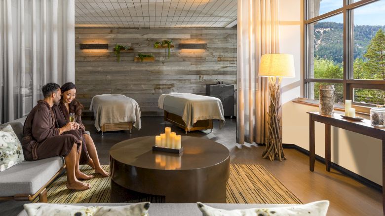 A cozy spa lounge with two people on a sofa, a round coffee table, warm lighting, candles, and two massage beds visible by a large window.
