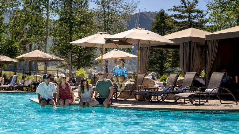 A group of people sits by a pool near lounge chairs and umbrellas, while staff or guests chat by the water in a resort setting.