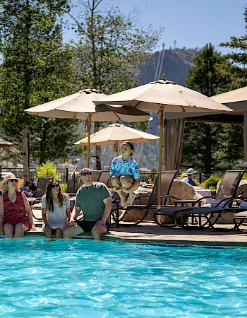 People sit and chat at a resort pool, with lounge chairs, umbrellas, and cabanas; trees and mountains in the background.