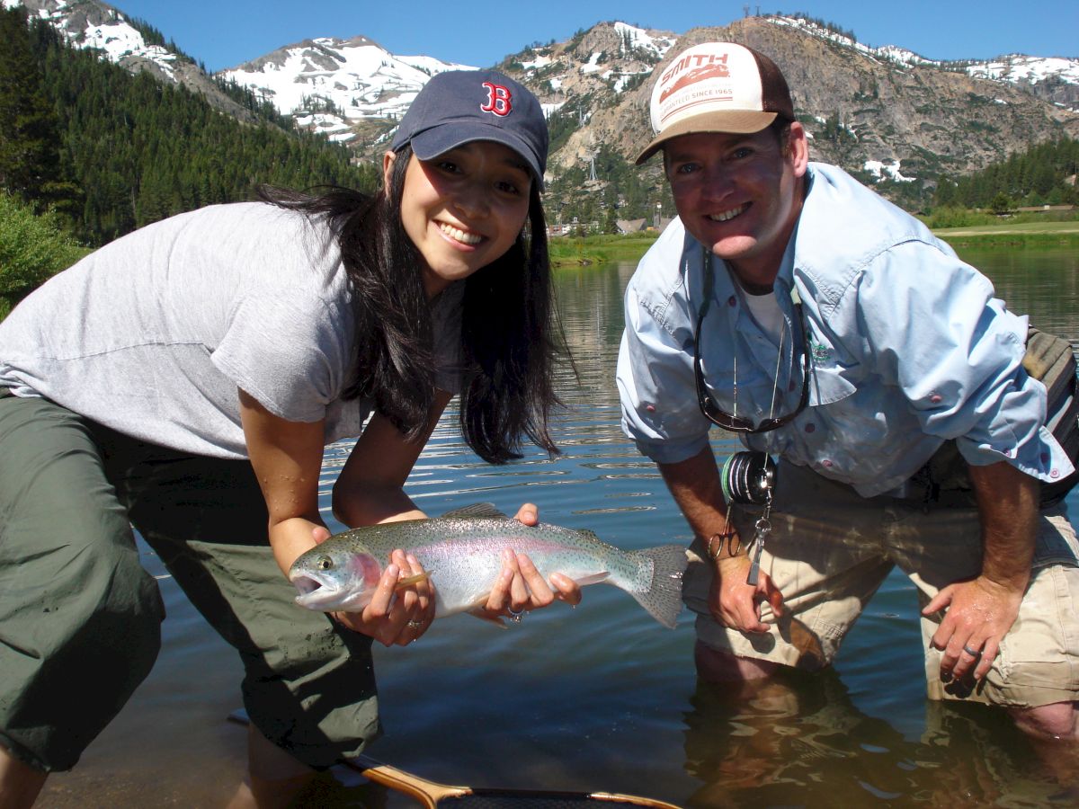 Two people standing in shallow water, one holding a fish, with mountains and snow in the background.