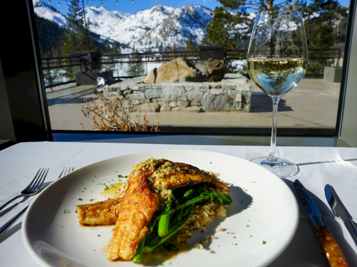 A plate of fish with greens and couscous on a table, accompanied by a glass of white wine, with a snowy mountain view outside the window.