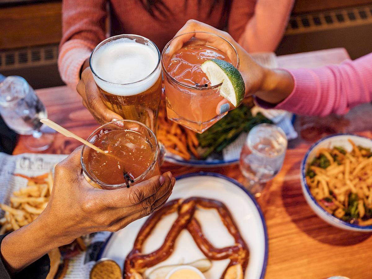 The image features three people toasting with drinks over a table laden with food, including a large soft pretzel, fries, and a bowl of noodles.