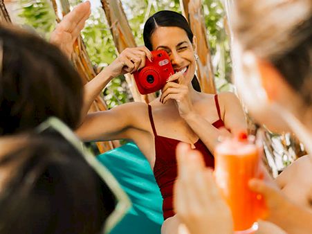 A group of friends at the beach or pool, posing for photos and toasting with drinks, sunny and cheerful vibes, one person holds a red camera.
