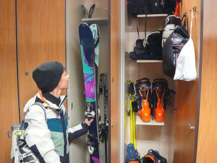 A person stands with ski gear next to open lockers containing skis, boots, and other equipment, possibly in a ski resort or rental shop.