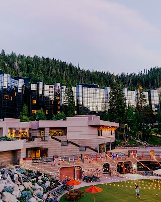 The image shows a scenic resort nestled in a forested area with modern buildings, a waterfall feature, string lights, and a lush green lawn, creating a tranquil atmosphere.