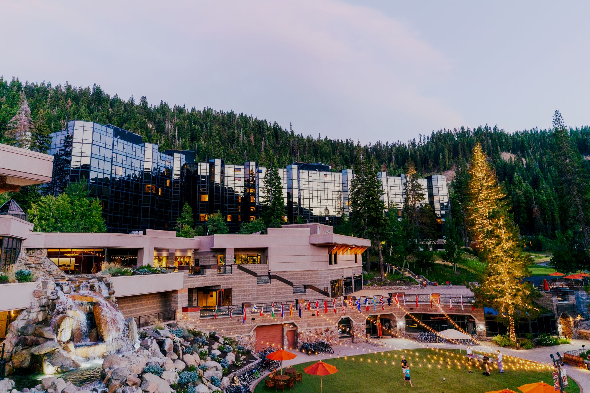 Modern buildings surrounded by lush trees at the edge of a forest, with a waterfall, lawn, and string lights in the foreground ending the sentence.