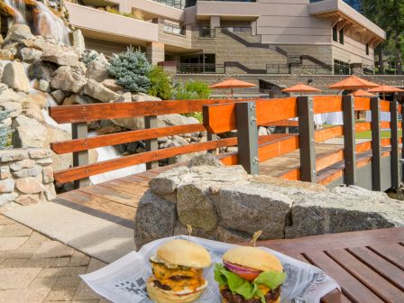Outdoor patio by a waterfall with grilled burgers on a table, orange umbrellas, and a modern lodge building in a pine forest backdrop.