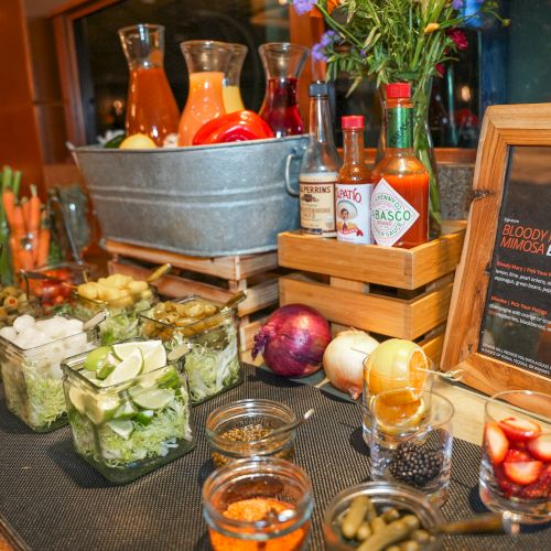 A bar setup for Bloody Mary & mimosas with mixers, bottles, garnishes, and a bucket of colorful sauces.