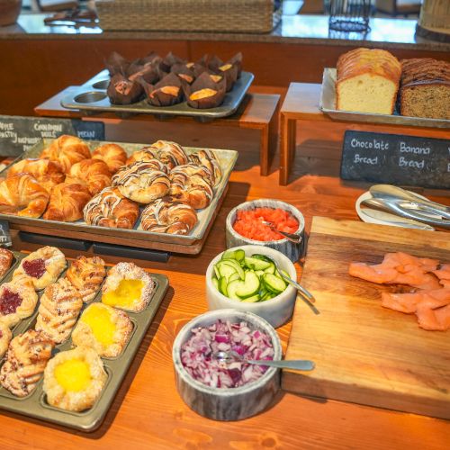 A breakfast buffet display with breads and pastries, plus sliced fruit like watermelon, cucumber, and pickled onions.