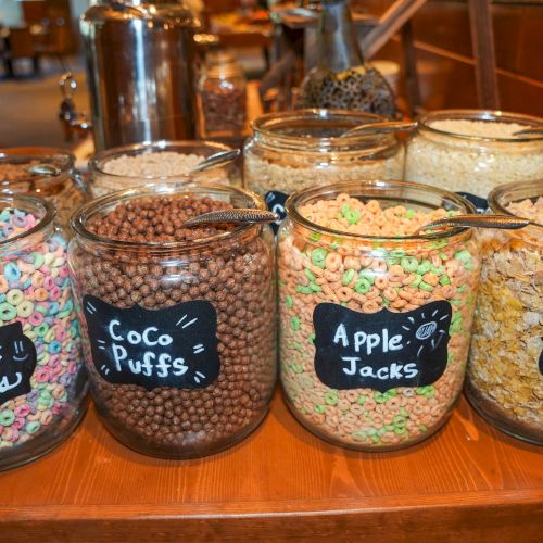 Glass jars of colorful cereal with chalk signs reading &ldquo;Apple Jacks,&rdquo; &ldquo;Frosted Flakes,&rdquo; &ldquo;Coco Puffs,&rdquo; and &ldquo;Froot Loops.&rdquo;