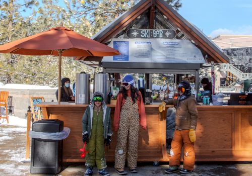 A small ski lodge with a wooden counter, two kids in snowsuits, and adults as staff under a peaked roof, with snow on the ground.