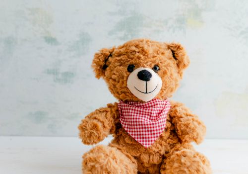 A cute plush teddy bear in a red plaid bandana sits on a light table against a pale blue wall.
