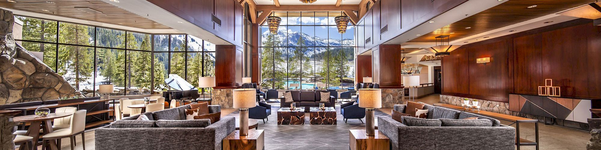A luxury lodge lounge with a grand chandelier, upholstered seating, and huge windows showing pine trees and distant mountains.