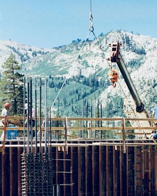 The image shows construction workers on a building site with mountains in the background, and a crane lifting materials.