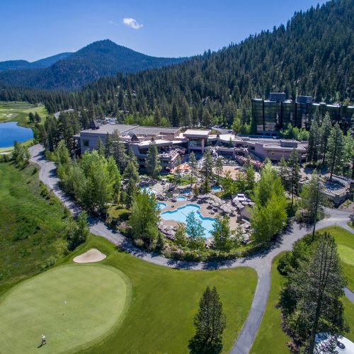 This image depicts a resort nestled in a mountainous area surrounded by lush greenery, with a pool, various buildings, and a golf course.