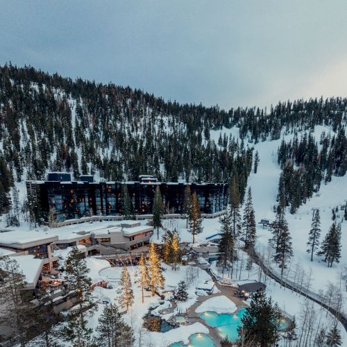 An aerial view of a snow-covered mountain resort with buildings, trees, and outdoor pools surrounded by a forested area and ski slopes in the background.