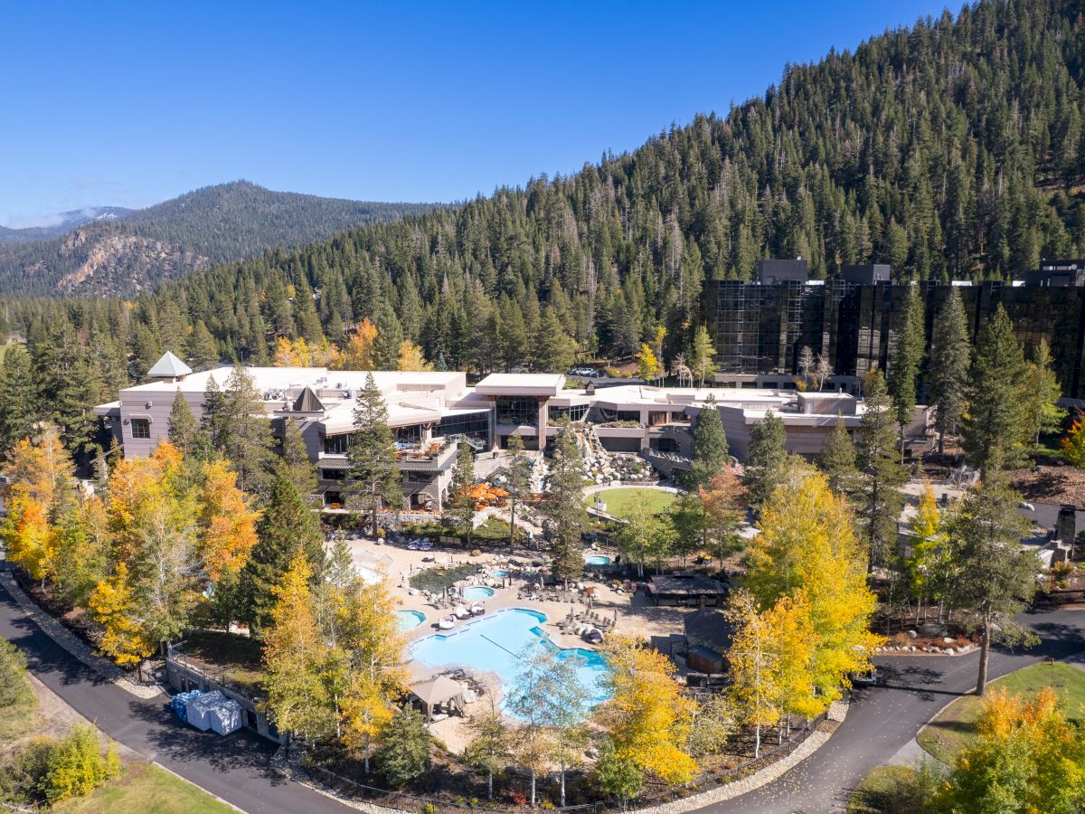 A mountain resort with a swimming pool and lodgings, surrounded by autumn trees and pine-covered hills under a blue sky.