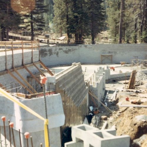 The image depicts a construction site with concrete walls, steel reinforcements, and various construction materials scattered around in a forested area.