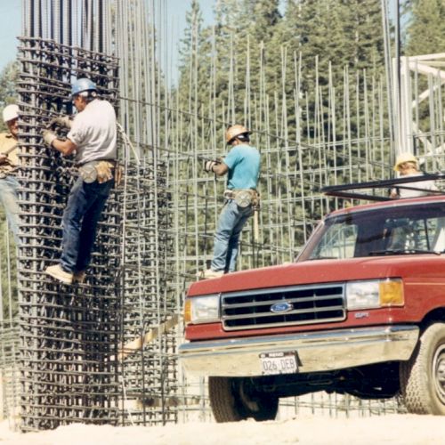 Construction workers are building a rebar structure with a red pickup truck nearby, surrounded by scaffolding and trees in the background, ending the sentence.