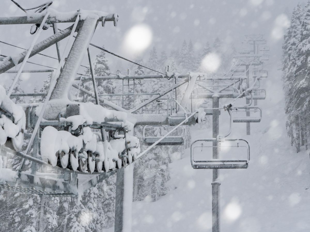 The image shows a snowy ski lift surrounded by a dense snowstorm. The lift and landscape are covered in snow, with tall trees in the background.