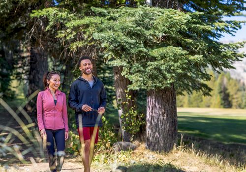 Two people are jogging on a sunny park path, smiling as they pass a large tree.
