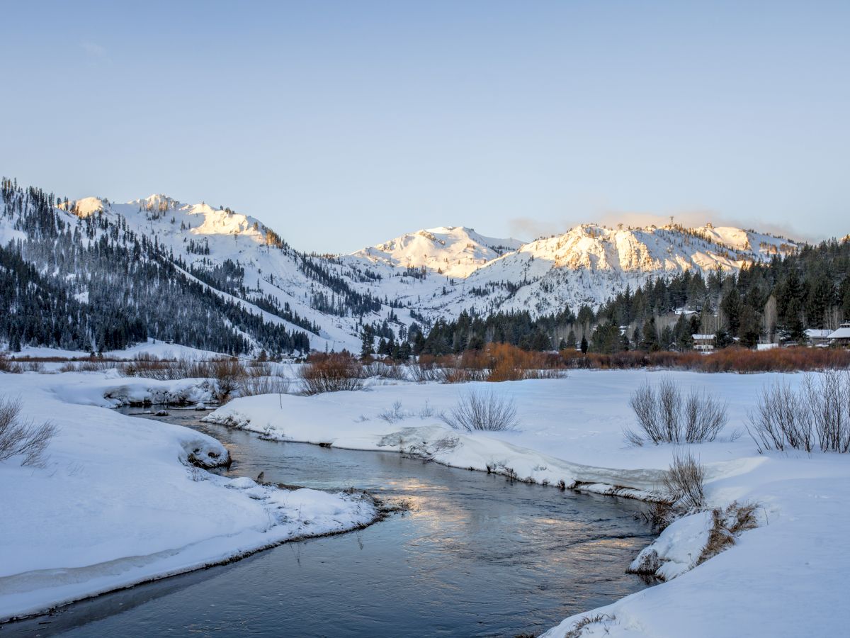 A snowy landscape features a serene river flowing through a valley, surrounded by snow-covered mountains and evergreen trees under a clear sky.