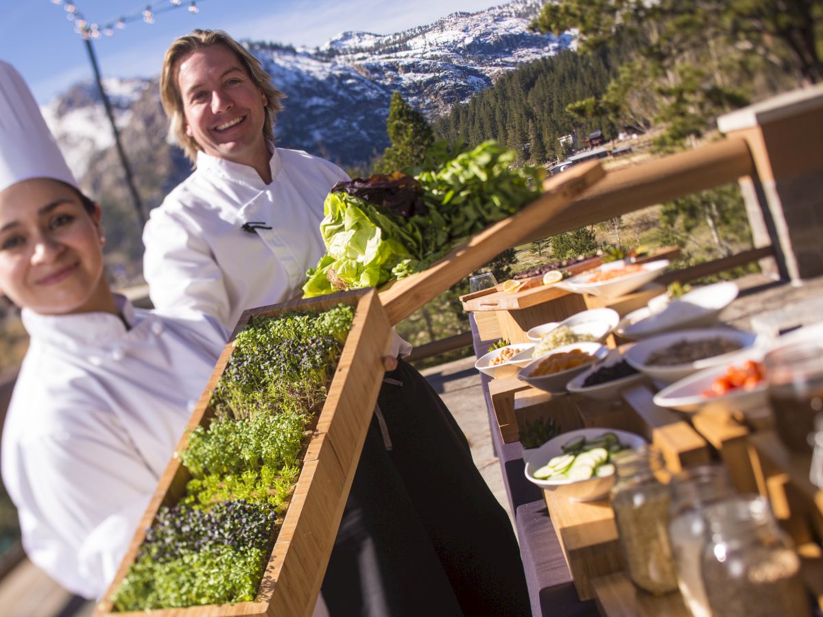 Two chefs outdoors, displaying a variety of fresh herbs and ingredients on a sunny day, with a snowy mountain landscape in the background.