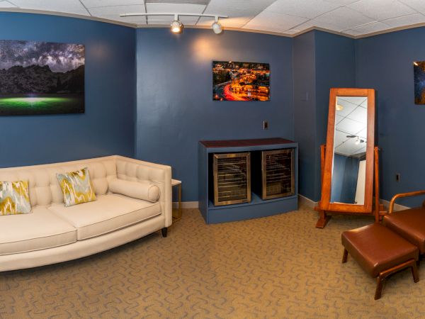 A small lounge room with a white sofa, leather chair, framed photos, a glass cabinet, patterned carpet, and a doorway mirror.