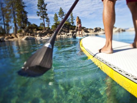 Two people paddleboarding on clear, calm water with trees and rocks in the background. The image focuses on one person's paddle and board.