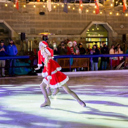 Two ice skaters dressed in festive Santa outfits perform on an ice rink, with a crowd watching them under string lights.