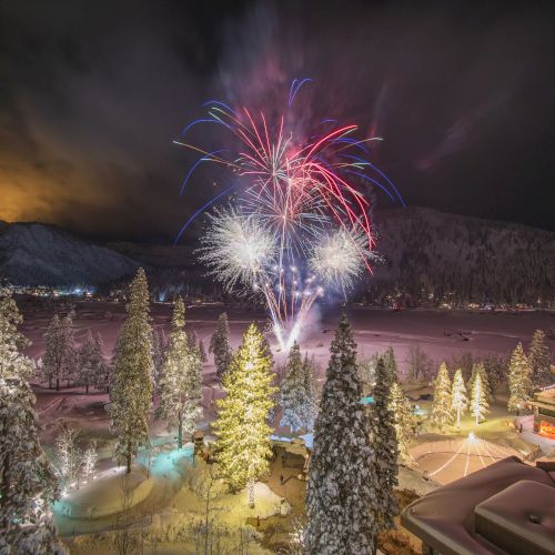 The image shows a snowy landscape with tall trees and buildings. Fireworks are exploding in the sky, illuminating the scene at night.