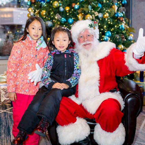 Two children are posing with Santa Claus in front of a decorated Christmas tree, with Santa making a waving gesture.