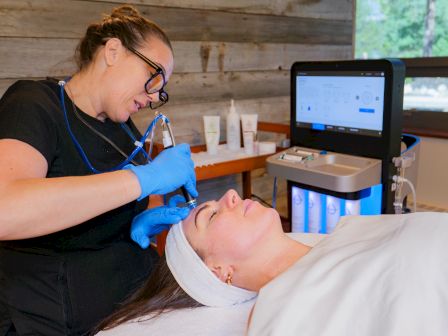An esthetician in gloves performs a facial treatment on a client while using a machine with a screen in a clinic.